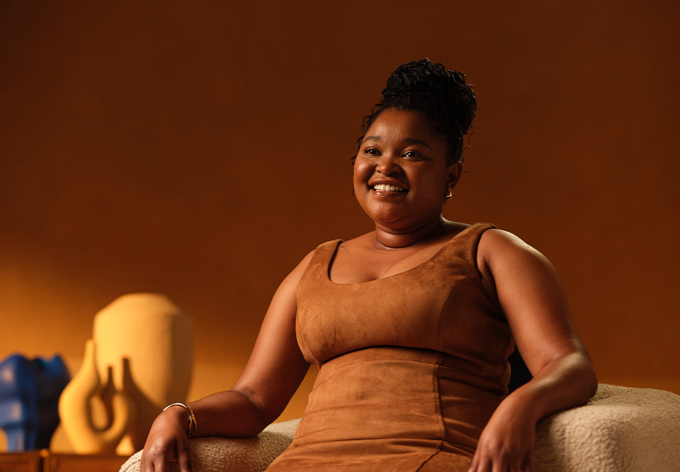 Lukhona, a woman with braided hair, wearing a brown sleeveless dress, sitting on a chair and smiling